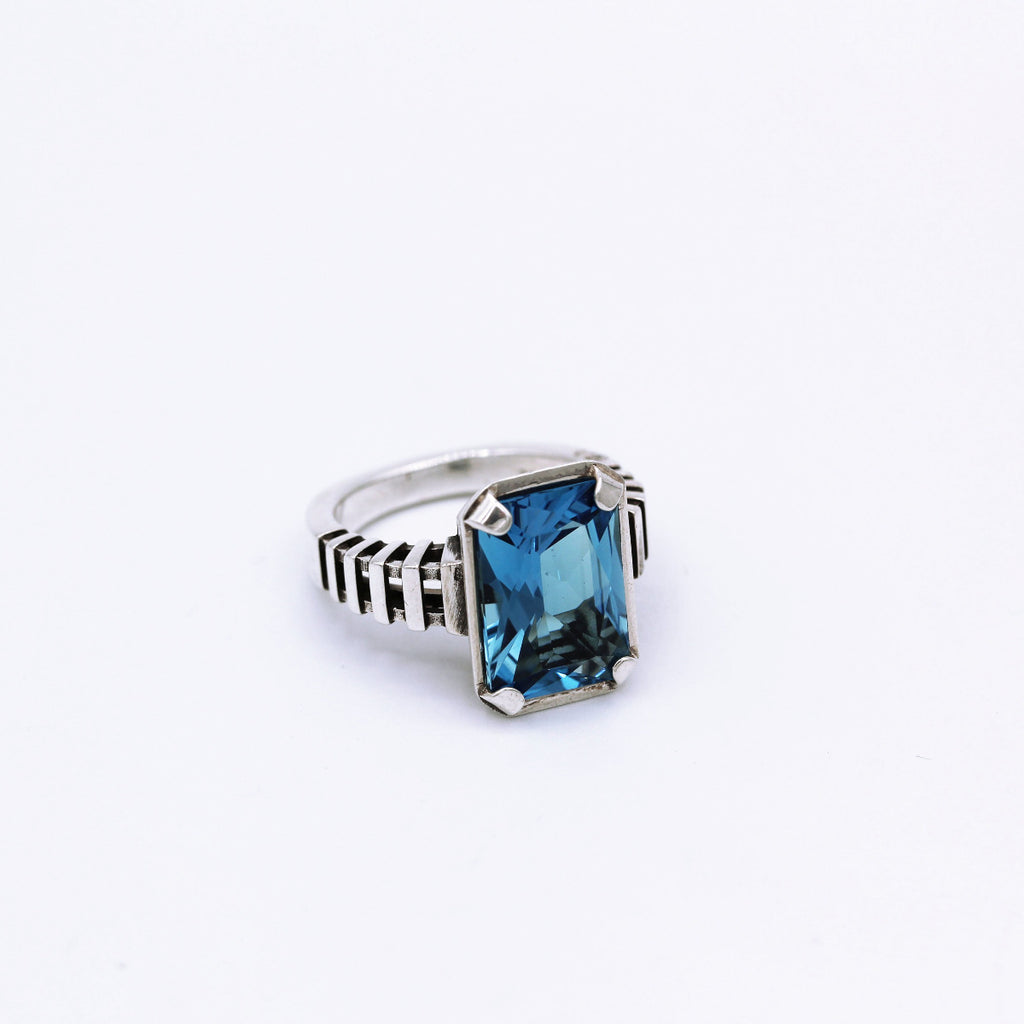 A sterling silver ring with an emerald cut blue topaz gemstone, featuring a minimalist design with intricate textures on the band.