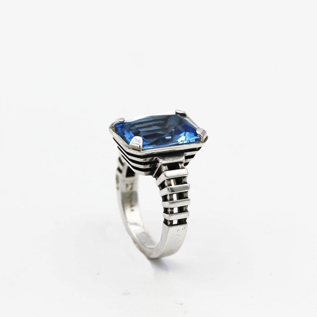A sterling silver ring with an emerald cut blue topaz gemstone, featuring a minimalist design with intricate textures on the band.