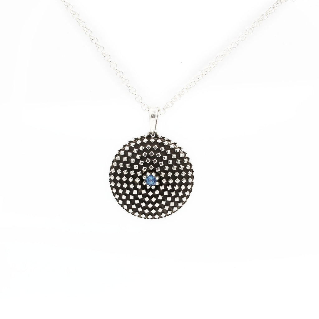 A silver pendant with a geometric diamond-cut design featuring a central blue topaz gemstone, suspended from a chain.